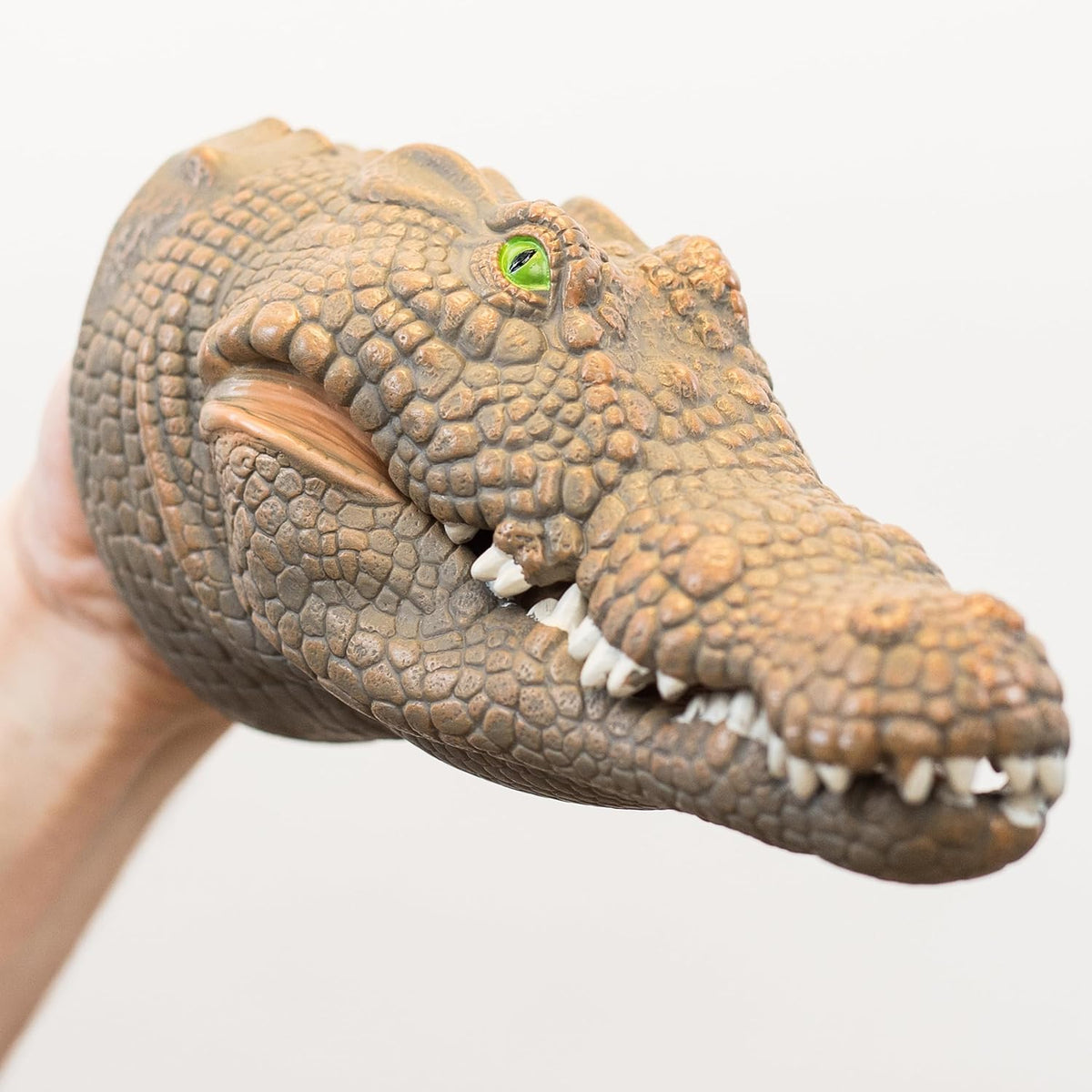 A realistic crocodile or alligator head hand puppet being held by a human hand against a plain light gray background.