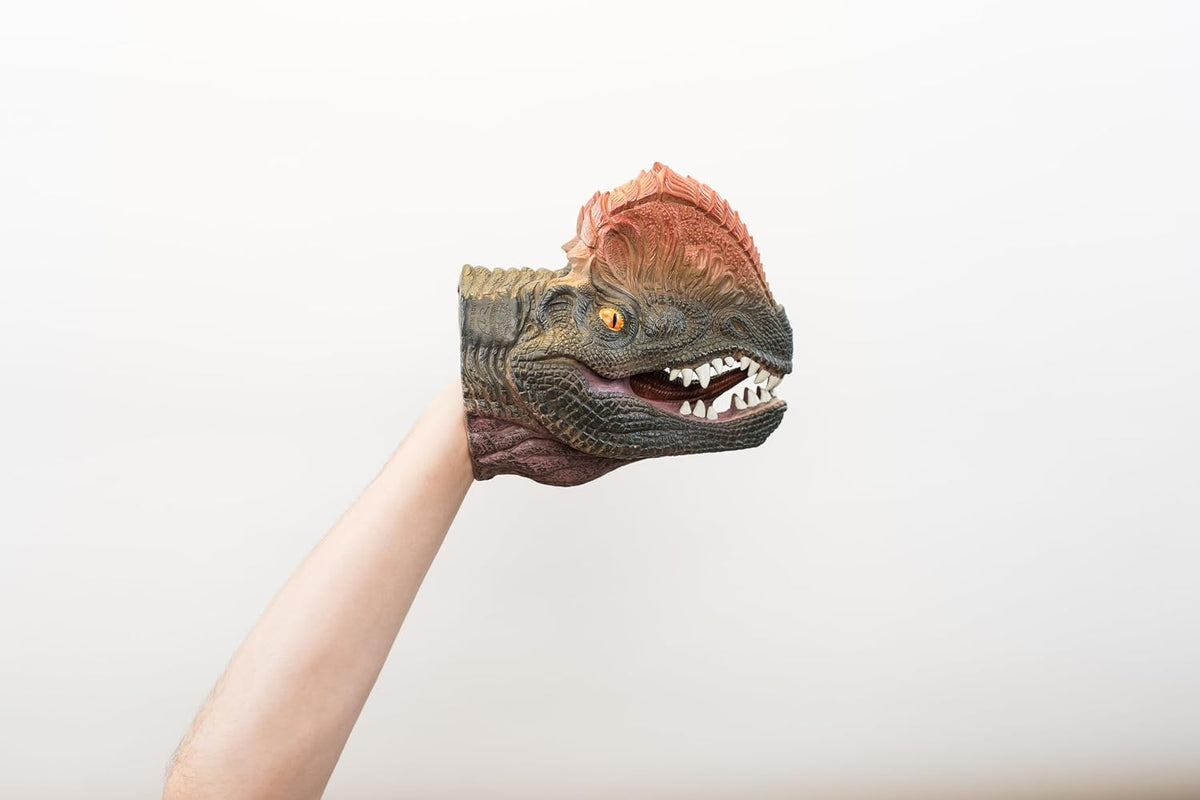 A dinosaur hand puppet with a textured green and brown body and a reddish-brown crest on its head, worn on a person's hand and held up against a plain light background.