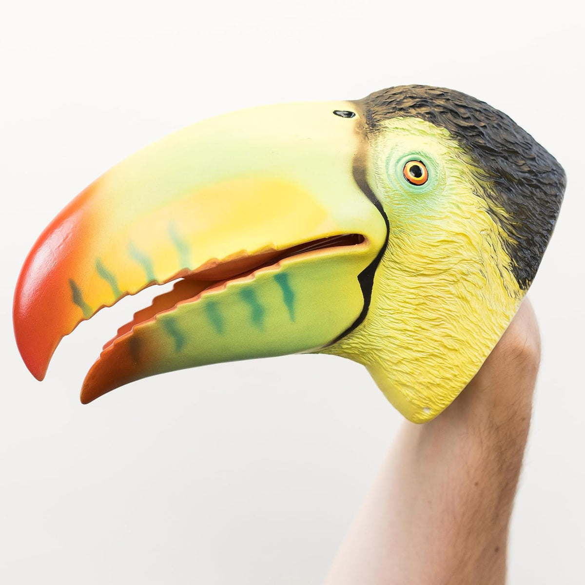 A hand wearing a realistic toucan head hand puppet mask, featuring a yellow face, green beak interior, orange-red tip, and black head, set against a plain white background.