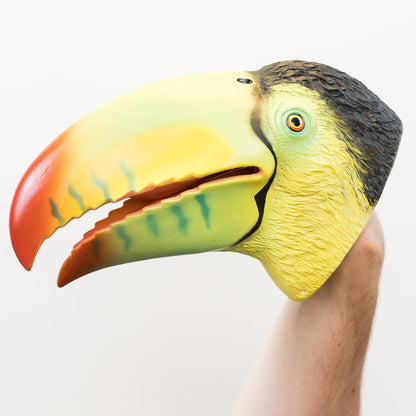 A hand wearing a realistic toucan head hand puppet mask, featuring a yellow face, green beak interior, orange-red tip, and black head, set against a plain white background.