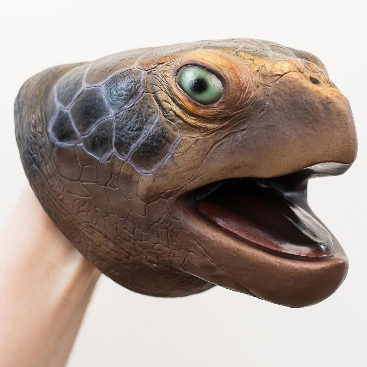A realistic turtle head hand puppet being held by a human hand, featuring detailed textured scales, a green eye, and an open mouth.
